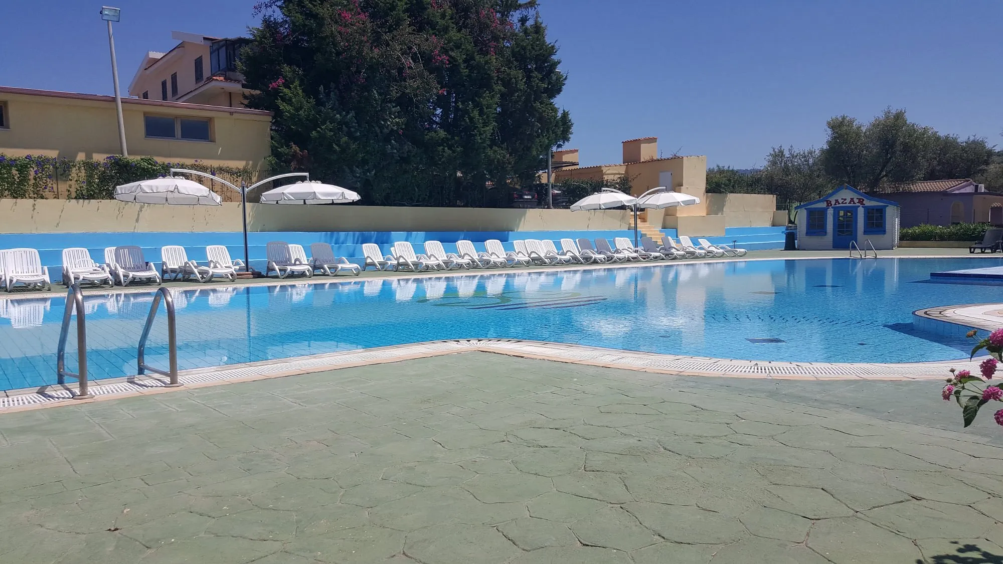 Grande piscina con lettini bianchi e chiosco bazar azzurro accanto agli edifici del resort