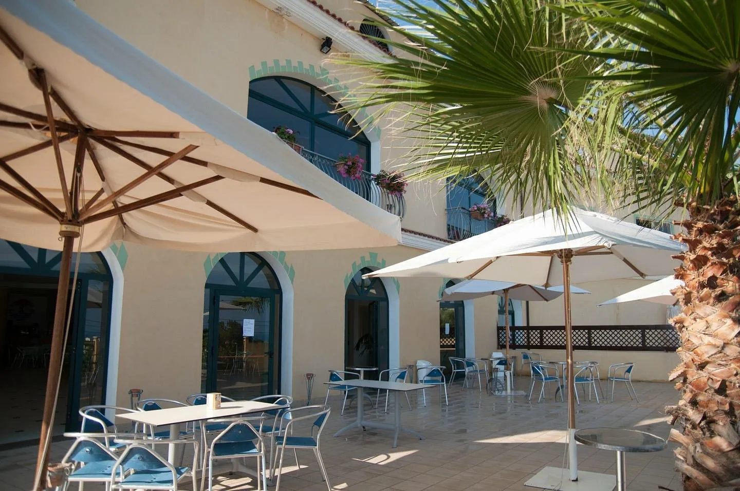 Terrazza esterna di ristorante con tavoli, sedie e ombrelloni bianchi accanto a palme