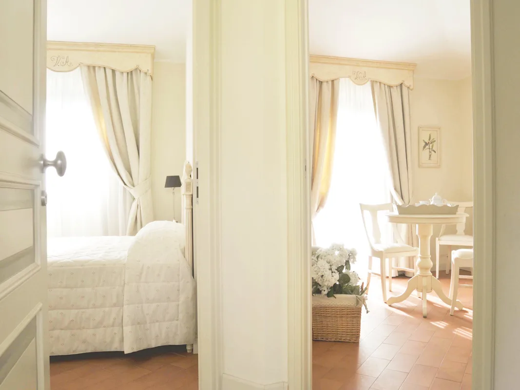Vista su camera con letto bianco e salottino con tavolo rotondo e fiori