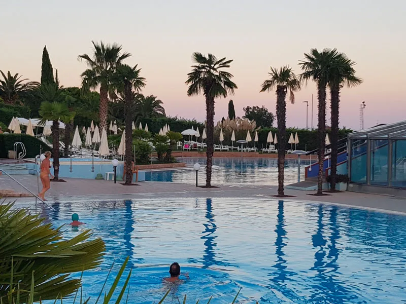 Piscina al tramonto con palme e bagnanti, cielo rosato