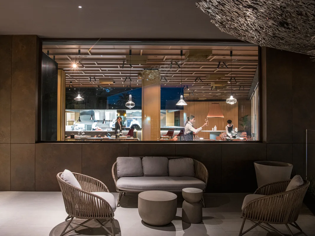 Lounge dell'hotel con poltrone in vimini e vista sulla cucina a vista del ristorante illuminata di sera