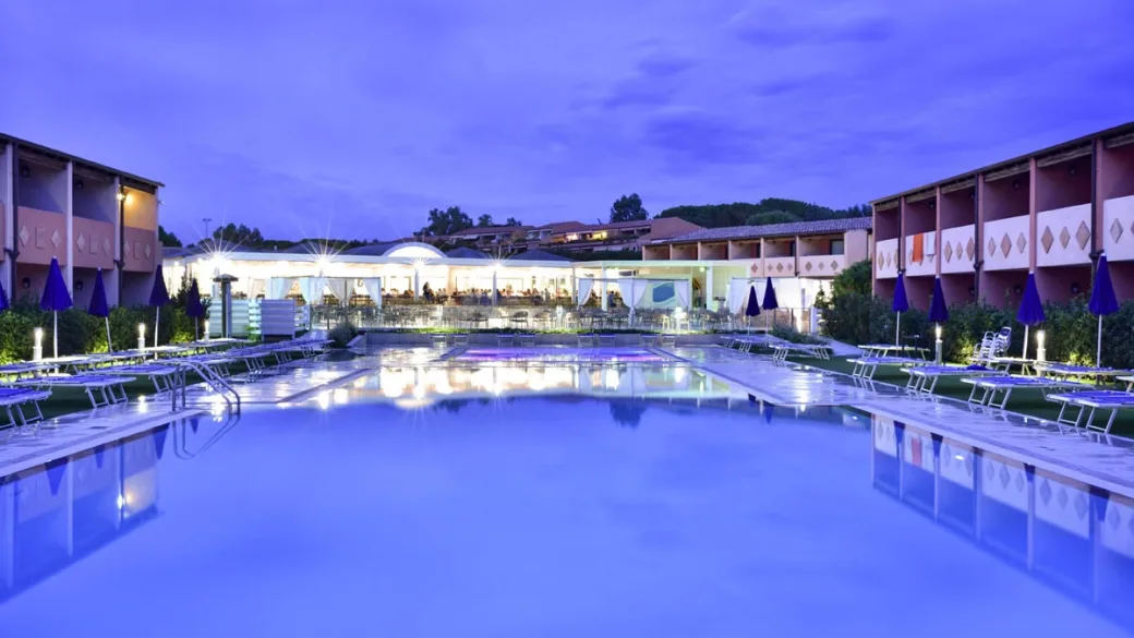 Piscina del resort al crepuscolo con ombrelloni blu e ristorante illuminato sullo sfondo
