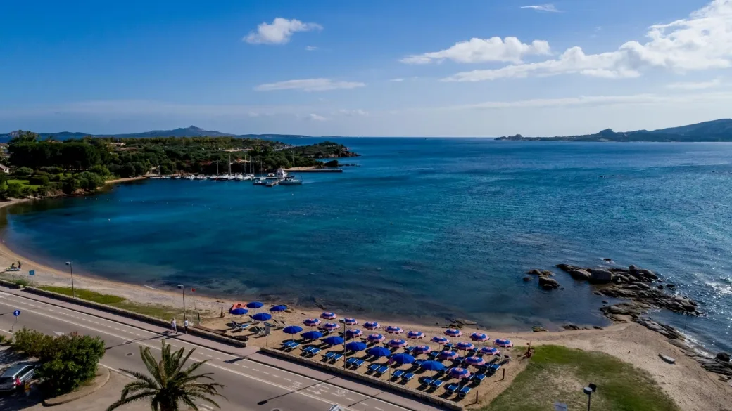 Panorama della spiaggia con ombrelloni blu, mare azzurro e porticciolo turistico