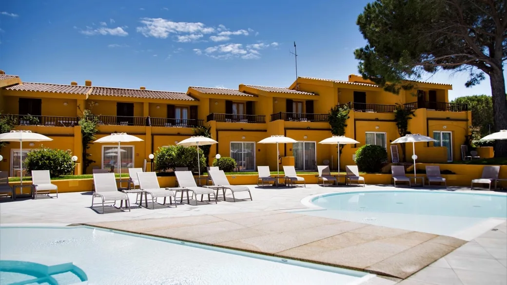 Area piscina con lettini, ombrelloni e edifici gialli del resort